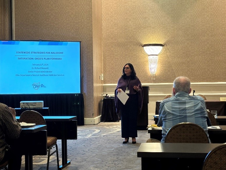 HMA Principal Teresa Garate, PhD presents at the 2024 Opioid & Fentanyl Abuse Management Forum sponsored by the World Conference Forum. She is standing at the front of a room next to a screen.